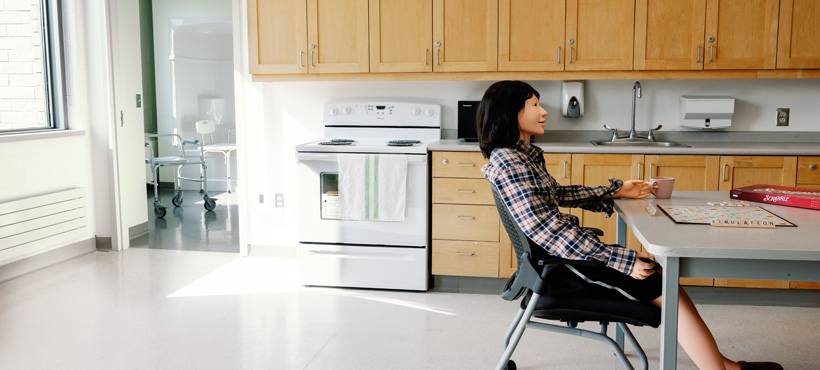 Sakura mannequin sits at dining table, holding a cup while playing a game of scrabble, with kitchen and bathroom partially visible in background