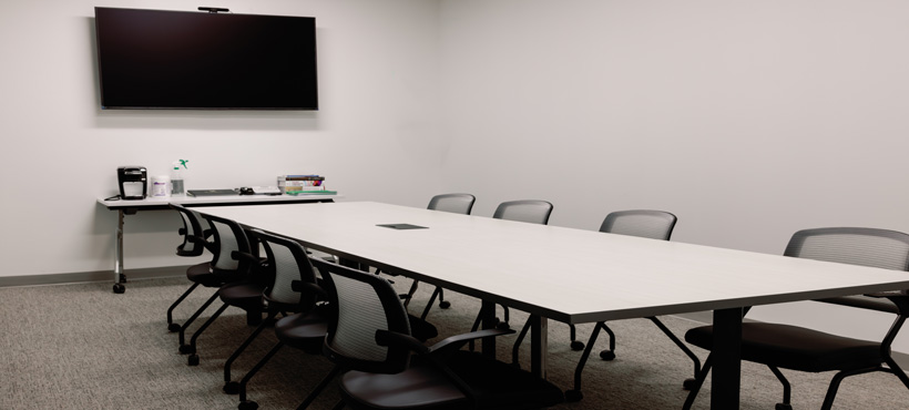 Debriefing space with long table surrounded by eight chairs, with wall-mounted large television and webcam to allow for video conferencing if desired