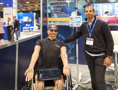 School of Health staff (seen on right side) stands next to Adolescent simulator 'Ares' (seen on left side) who is sporting some Mohawk College gear, seated in a wheelchair with an intravenous transfusion running