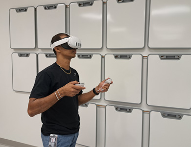 Young male staff member wearing virtual reality headset holding controllers in each hand, standing in front of wall with numerous mounted small whiteboards