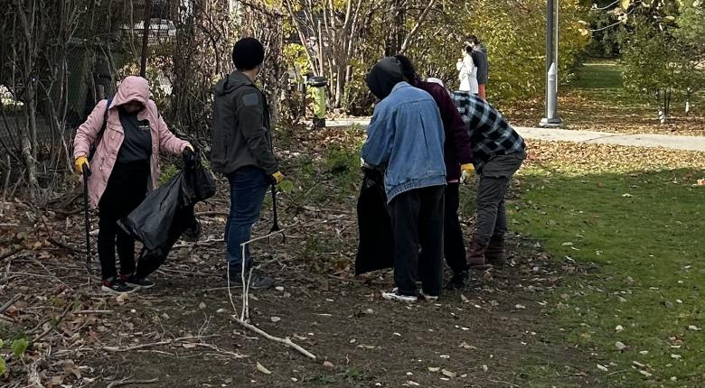 Four students collecting litter and placing it in a garbage bag during the Fall season. Two more students are also participating in the clean-up in the background.