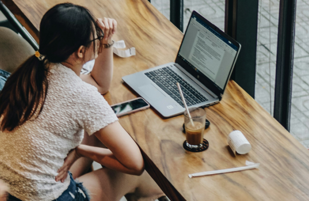 An image of a woman sitting at a table in a coffee shop, looking intently at her laptop.