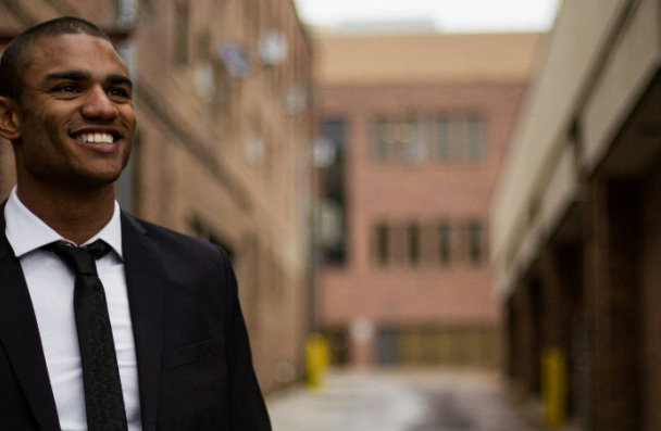 A darker skinned man in a suit with a smile on his face, standing in an urban area