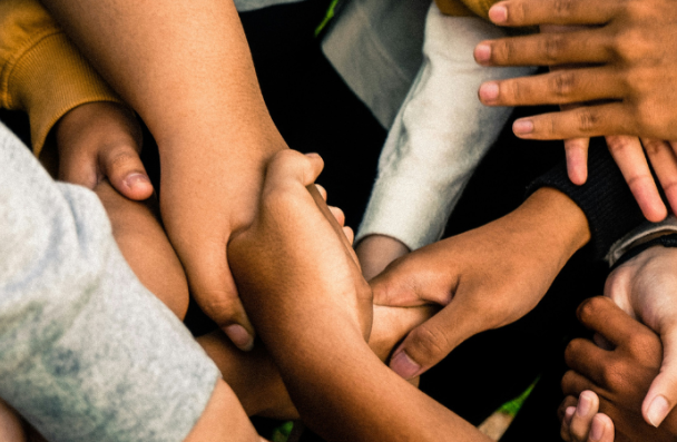 An image of a group of hands, all reach across each other to hold another hand.  The hands are different sizes and skin tones.